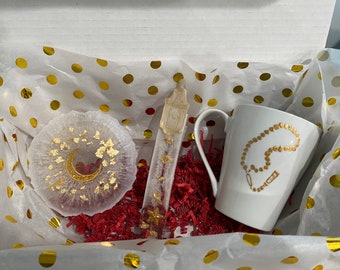 May include: A gift box filled with a white mug with a gold chain design, a white resin coaster with a gold crescent moon and gold flakes, and a clear glass tube with a gold lantern charm.