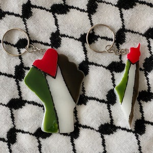 May include: Two keychains, one shaped like a fist with a red heart and the other shaped like the outline of Palestine, both with the colors of the Palestinian flag.