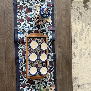 May include: A table setting with a floral runner with a navy border. A wooden tray holds six small cups with white interiors and blue rims. A patterned teapot and a book are also present on the table.