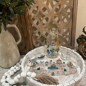May include: A white wooden tray with a hand-painted scene of a city with gold and blue accents. The tray includes four clear glass cups and a glass pitcher with a gold tassel.