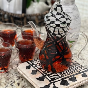 May include: A black and white patterned glass teapot with a lid, filled with red tea, sitting on a white wooden tray with a black and white patterned design.