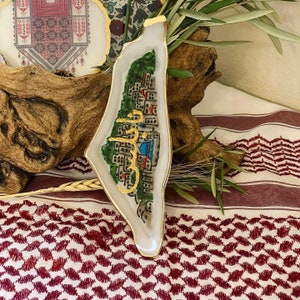 May include: A white resin tray shaped like the outline of Palestine with a gold trim. The tray features a painted cityscape with green trees and a blue mosque. The Arabic word "Palestine" is written in gold on the tray.