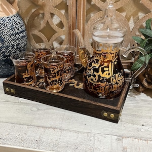 May include: A glass tea set with a dark brown tray. The set includes a clear glass teapot with gold calligraphy, four small glasses, and a tray. The glasses and teapot are filled with tea. The tray has brass corner accents.