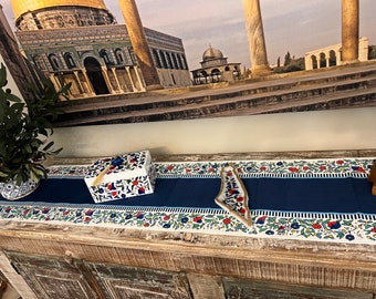 May include: A blue and white table runner with a floral pattern. The runner is on a wooden table with a white box and a small plate on it. The background is a picture of a mosque.