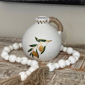 May include: White ceramic vase with a rope handle and a green olive branch design. The vase is sitting on a wooden surface with a white beaded garland with a brown tassel.