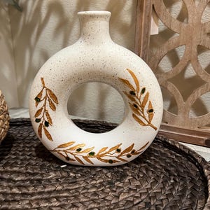 May include: White ceramic vase with a ring-shaped opening and gold and green leaf designs. The vase is sitting on a woven brown surface.