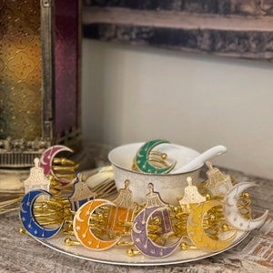May include: A set of eight gold-coloured napkin rings featuring a crescent moon and lantern design. The rings are decorated with stars and come in a variety of colours, including blue, orange, yellow, purple, and white.