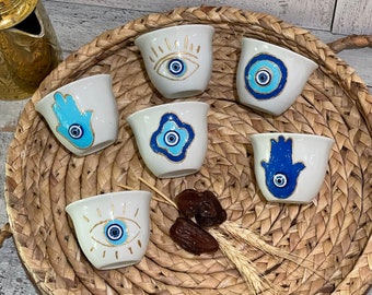 May include: Six white ceramic coffee cups with blue and gold designs. The designs include an evil eye, a hamsa hand, and a flower. The cups are arranged in a circle on a woven tray.