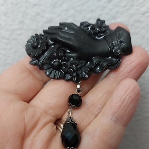 May include: Black cameo brooch featuring a hand holding a flower, with a dangling black crystal drop.