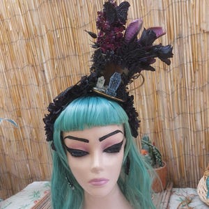 May include: A black headpiece with dark purple and black floral accents, a miniature coffin, and a small hat. The headpiece is worn by a mannequin with turquoise hair and dramatic eye makeup. The background is a bamboo screen.