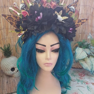 May include: A decorative headpiece featuring a black floral base adorned with leopard print accents, stylized plant elements, and small, colorful details. The headpiece sits atop a mannequin head with blue hair.