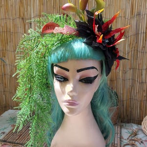 May include: A decorative headpiece featuring artificial green fern fronds, red-tipped Venus flytrap plants, and a large black flower with orange and red accents. The headpiece is displayed on a mannequin head with light teal hair and dark makeup.