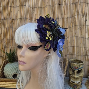 May include: A hair accessory featuring a large dark purple dahlia flower, accented with smaller light blue orchid petals, green and yellow Venus flytrap motifs, and dark spiky thistle elements, worn on a mannequin head with white hair.