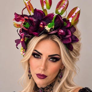 May include: A decorative headband featuring dark purple orchid flowers, bright green Venus flytrap plants, and a small bird skull. The accessory is adorned with vibrant green moss accents and is worn with ornate dangling earrings and a black lace choker.