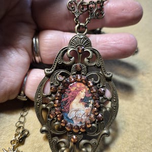 May include: Antique bronze-toned pendant necklace featuring a central oval image of a woman with flowing red hair. The pendant is adorned with brown crystals and intricate filigree detailing. A smaller sunburst charm hangs from the chain.