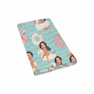May include: A teal blue beach towel with a repeating pattern of people's faces on various inflatable pool floats, including a unicorn, a swan, and a pink flamingo. The floats are surrounded by pink and white donuts with sprinkles.