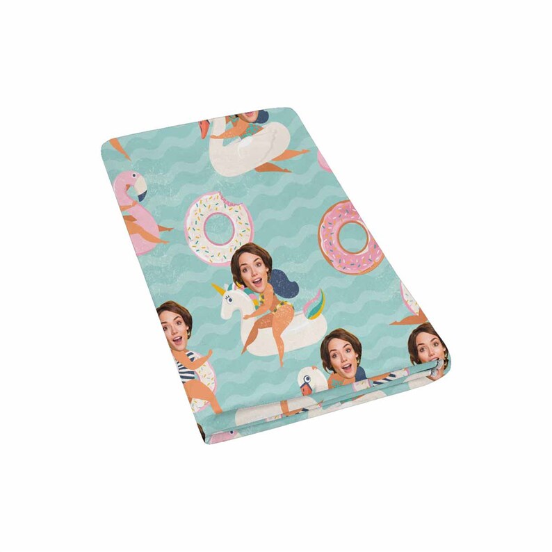 May include: A teal blue beach towel with a repeating pattern of people's faces on various inflatable pool floats, including a unicorn, a swan, and a pink flamingo. The floats are surrounded by pink and white donuts with sprinkles.