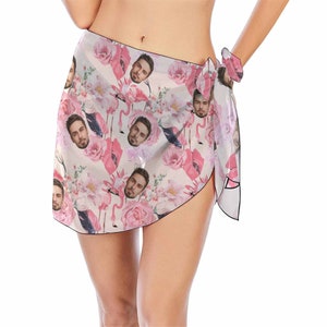 Pu&ograve; includere: Un pareo bianco con un motivo floreale rosa e un'immagine ripetuta del viso di un uomo. Il pareo &egrave; legato in vita e ha un bordo con frange.