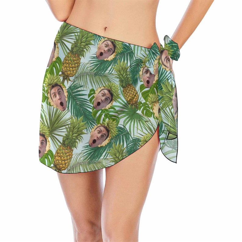 Pu&ograve; includere: Un pareo verde e bianco con una stampa tropicale con ananas e volti. Il pareo &egrave; legato in vita e ha un bordo con frange.