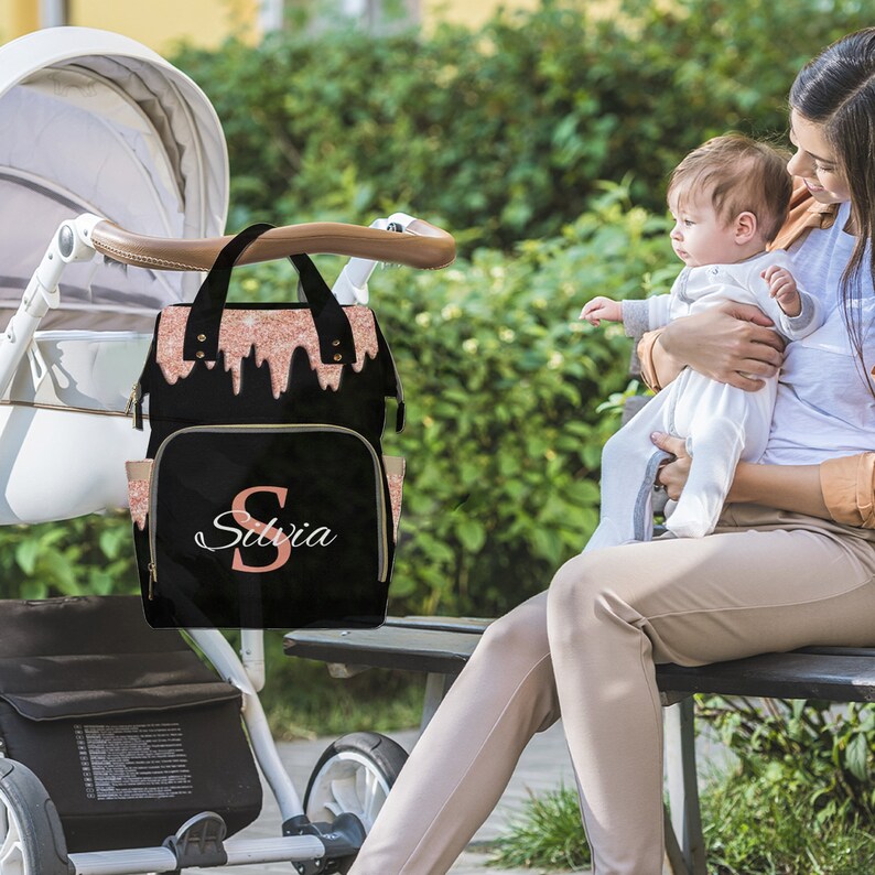 May include: A black diaper bag with a pink glitter design and the name "Silvia" in white lettering. The bag has a zippered pocket on the front and two side pockets. The bag is hanging on a stroller.