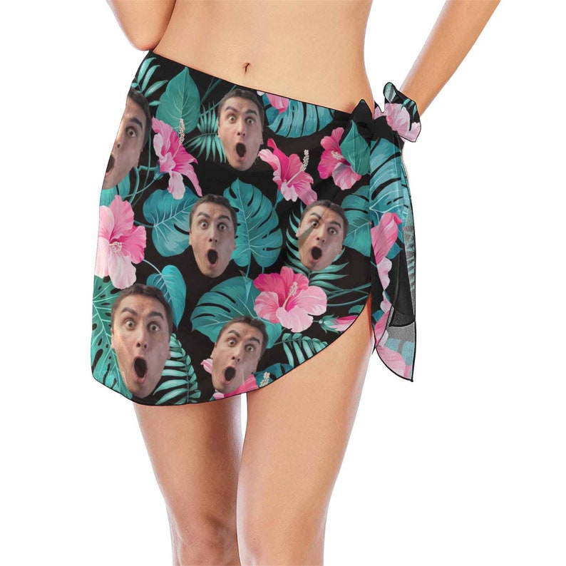 Pu&ograve; includere: Un pareo nero con una stampa tropicale con fiori rosa e foglie verdi. La stampa include anche pi&ugrave; immagini del viso di una persona.