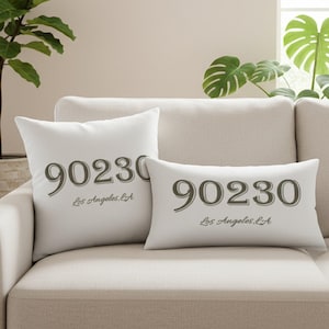 May include: Two white throw pillows on a beige sofa. The pillows display the number "90230" in a dark gray font, with "Los Angeles, CA" written below. One pillow is square, the other is rectangular.