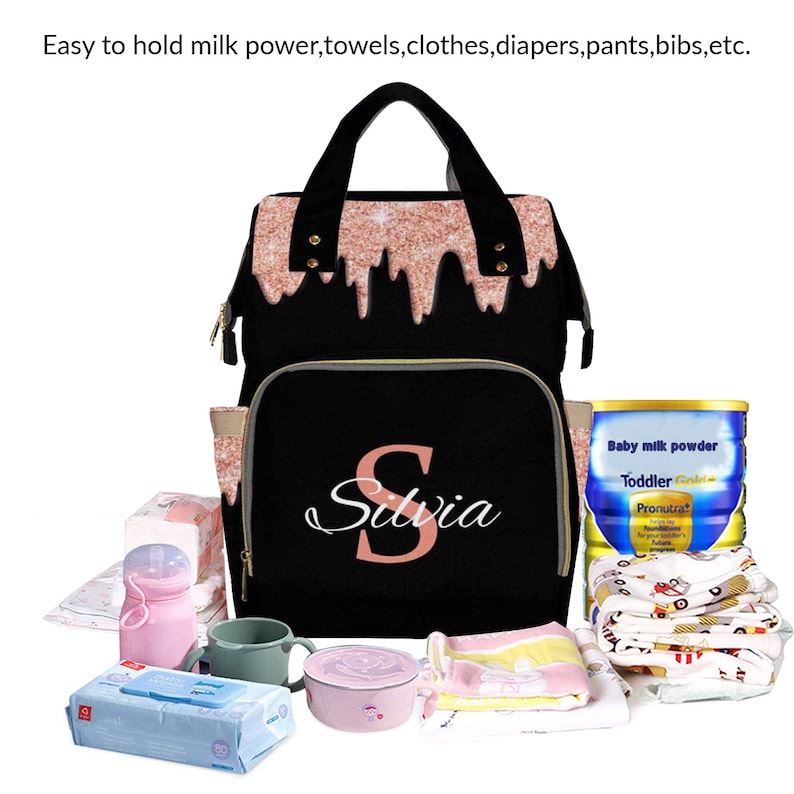 May include: A black diaper bag with a pink glitter design and the name "Silvia" printed on the front. The bag is filled with baby supplies, including a bottle of baby formula, wipes, and a pink sippy cup.