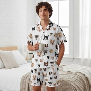 K&ouml;nnte beinhalten: Wei&szlig;es Pyjama-Set mit einem sich wiederholenden Muster verschiedener Hunderassen. Das kurz&auml;rmelige Hemd mit Kn&ouml;pfen und die passenden Shorts zeigen eine Vielzahl von Hundegesichtern in Braun-, Schwarz- und Wei&szlig;t&ouml;nen. Die Person h&auml;lt eine Tasse.