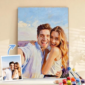 May include: A painted canvas depicts a smiling couple embracing, set against a lavender field and a blue sky. The painting has visible brushstrokes, and a smaller image of the couple is shown below. Paintbrushes and paint pots are visible.