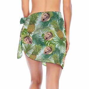 Pu&ograve; includere: Un pareo azzurro chiaro con una stampa tropicale di ananas e foglie di palma. La stampa include anche un'immagine ripetuta di un viso umano.