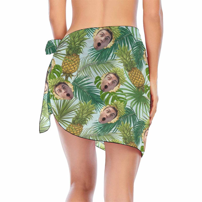Pu&ograve; includere: Un pareo azzurro chiaro con una stampa tropicale di ananas e foglie di palma. La stampa include anche un'immagine ripetuta di un viso umano.