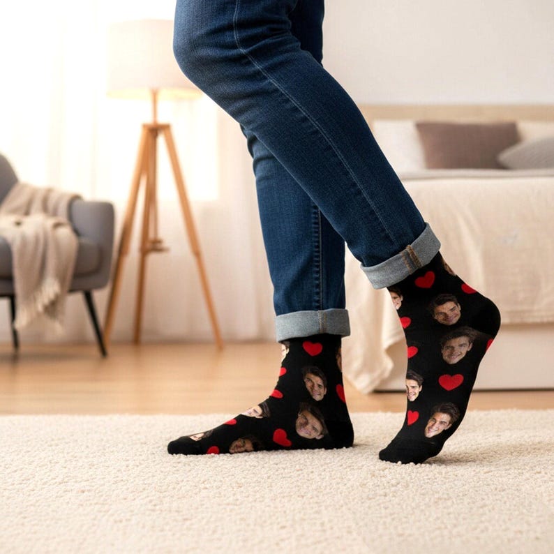May include: Black socks featuring a repeating pattern of faces and red hearts. The socks are worn with blue jeans. The socks are visible from the ankles down, with the person standing on a white carpet.