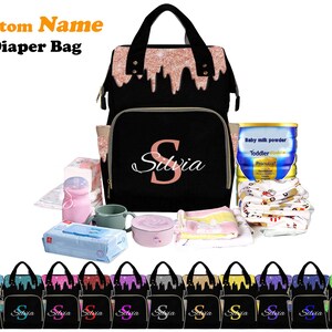 May include: A black diaper bag with a pink glitter design and a pink "S" monogram. The bag is shown with various baby items, including a bottle of baby formula, a container of baby wipes, a sippy cup, and a baby blanket.