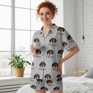 K&ouml;nnte beinhalten: Hellgraues Pyjama-Set mit einem sich wiederholenden Muster aus Dackelgesichtern und dem Wort "Olions". Das kurz&auml;rmelige Hemd mit Kn&ouml;pfen und die passenden Shorts sind aus weichem Stoff. Die Person im Bild h&auml;lt eine wei&szlig;e Tasse.