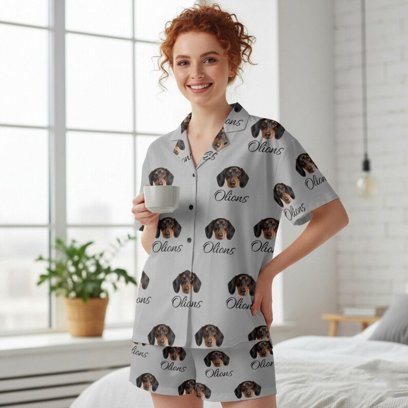 K&ouml;nnte beinhalten: Hellgraues Pyjama-Set mit einem sich wiederholenden Muster aus Dackelgesichtern und dem Wort "Olions". Das kurz&auml;rmelige Hemd mit Kn&ouml;pfen und die passenden Shorts sind aus weichem Stoff. Die Person im Bild h&auml;lt eine wei&szlig;e Tasse.