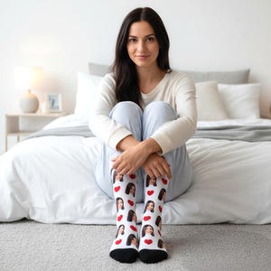 May include: White socks with a black toe and heel, featuring a repeating pattern of red hearts and a person's face. The socks are worn by a person sitting on a bed, wearing a white long-sleeve shirt and blue striped pants.