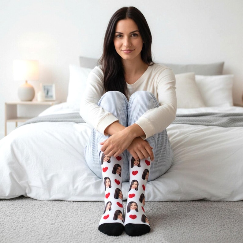 May include: White socks with a black toe and heel, featuring a repeating pattern of red hearts and a person's face. The socks are worn by a person sitting on a bed, wearing a white long-sleeve shirt and blue striped pants.