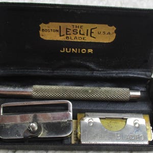 May include: A black leather case with a gold inscription that reads "The Boston Leslie Blade Junior" and contains a silver razor blade holder, a silver blade sharpener, and a pack of razor blades with the text "Leslie Blades".