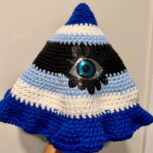 May include: A pointed crochet hat featuring horizontal stripes in royal blue, white, and light blue. The center of the hat displays a large, decorative eye patch embellished with black and silver sequins and a bright blue iris in the middle.