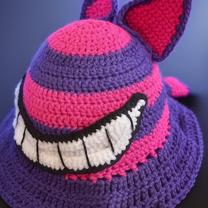 May include: A handmade crochet hat in purple, pink, and white, featuring a wide, toothy grin and pointed ears. The hat has alternating stripes of pink and purple, with a black outline around the grin and ears. The hat is a whimsical accessory.