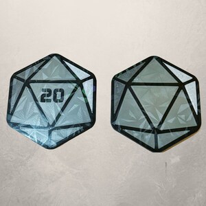 May include: A pair of 20-sided dice with a gray and black geometric design. The dice are made of a clear material with a textured surface.