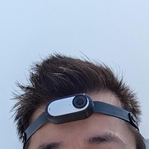 May include: A black head-mounted camera with a silver front panel is secured to a person's forehead with a black strap. The camera is centered on the forehead, and the strap is fastened around the head. The background is a light blue sky.