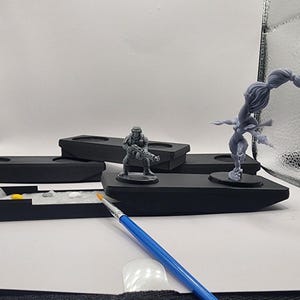 May include: A collection of miniature figurines, including a soldier and a female warrior, displayed on black platforms. A paintbrush and paint tray with various colors are also present, suggesting a hobby or craft activity.