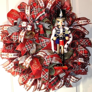 May include: A red, green, and white plaid Christmas wreath with a nutcracker holding drumsticks. The wreath is decorated with red and white ribbon, and the nutcracker is wearing a blue and gold uniform.