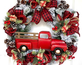 May include: A red truck Christmas wreath with a red and black buffalo plaid ribbon, green pine branches, and white flocked pine needles.