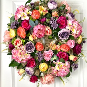 May include: A circular floral wreath featuring a dense arrangement of artificial roses, peonies, and hydrangeas in shades of pink, purple, orange, and yellow, interspersed with natural-toned burlap mesh ribbons and green foliage.