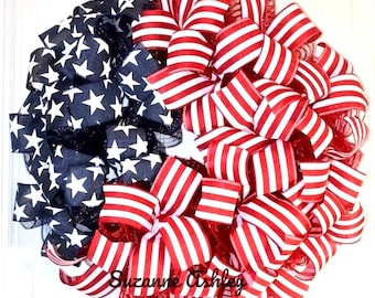 May include: A patriotic wreath featuring a design of the American flag. The wreath is divided into sections of navy blue with white stars and red and white striped ribbons. The text "Suzanne Ashley Designs" is visible.