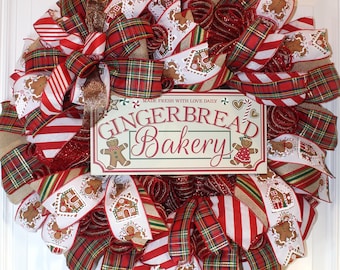 Puede incluir: Una corona navideña decorada con cinta de cuadros rojos y blancos, cinta blanca con hombres de jengibre y casas, y cinta de arpillera. La corona también tiene un letrero que dice "Gingerbread Bakery" con el texto "Made Fresh With Love Daily".