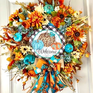 May include: A fall-themed wreath with orange, yellow, and turquoise flowers, leaves, and pumpkins. A sign in the center reads "Welcome" with a black and white checkered background.