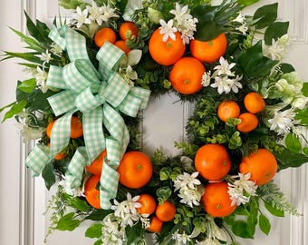 Può includere: Una ghirlanda di verde, fiori bianchi e frutta arancione è decorata con un fiocco di Vichy verde e bianco.